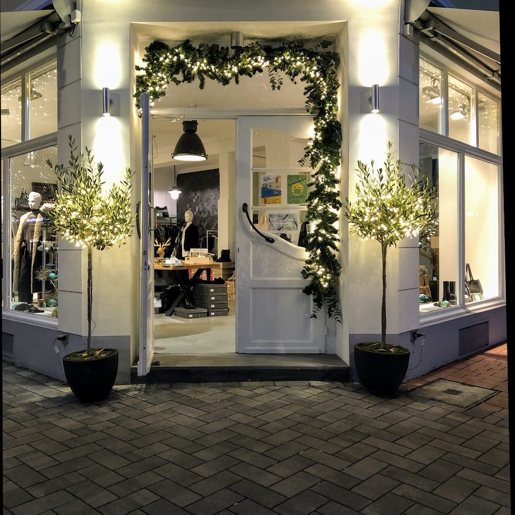 Der beleuchtete Eingang der Coccinella Boutique mit Schaufenstern und Olivenbäumen in Troisdorf
